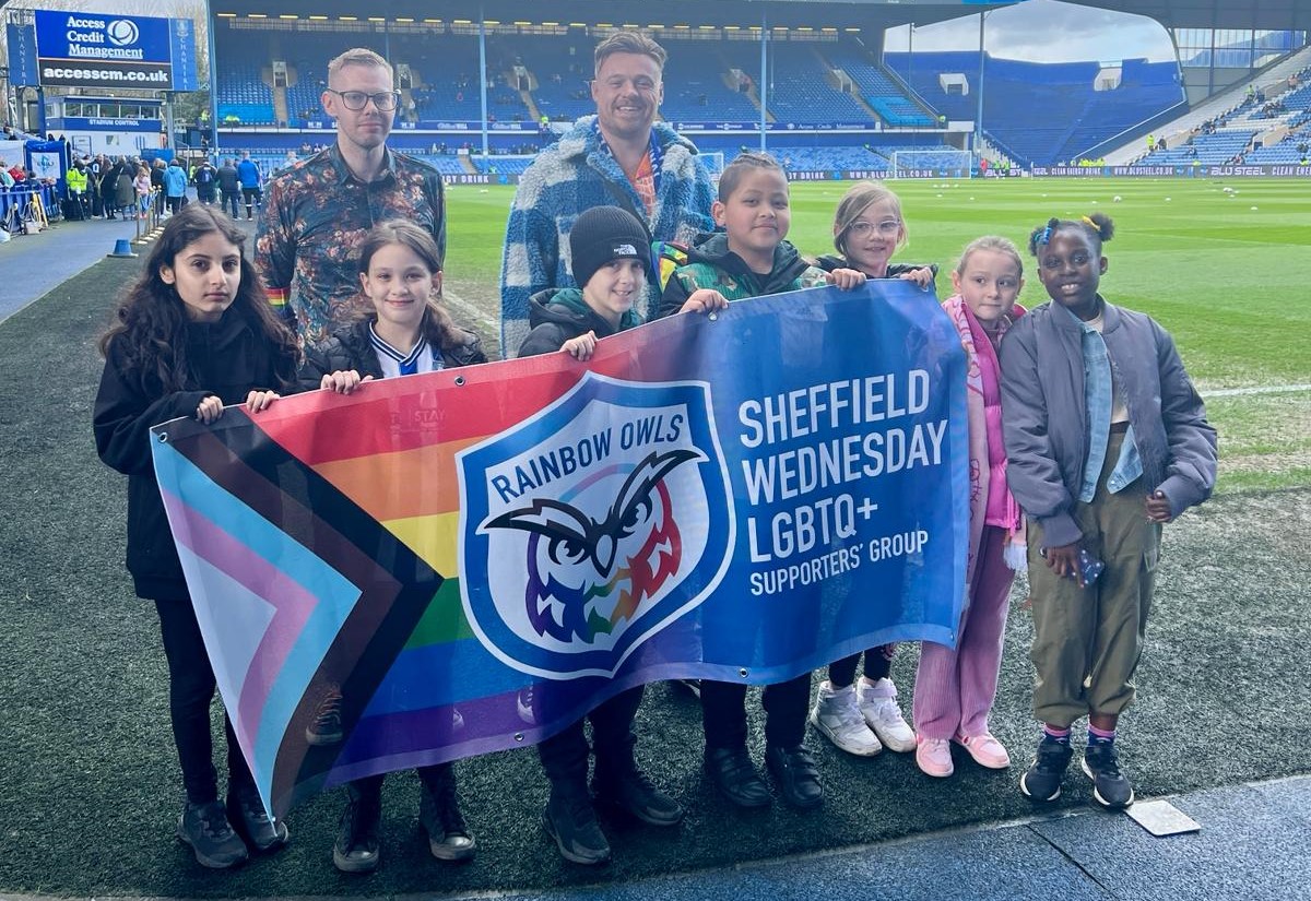 Rainbow Owls members holding the group banner with school children from Oasis Academy Watermead at Hillsborough Stadium before a designated Football v Transphobia matchday on Friday 29 March.